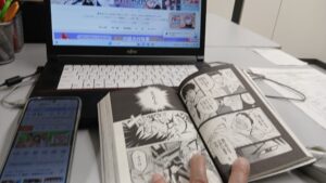 パソコンとスマートフォンで漫画の配信先を調べながら紙の漫画を読むイメージ